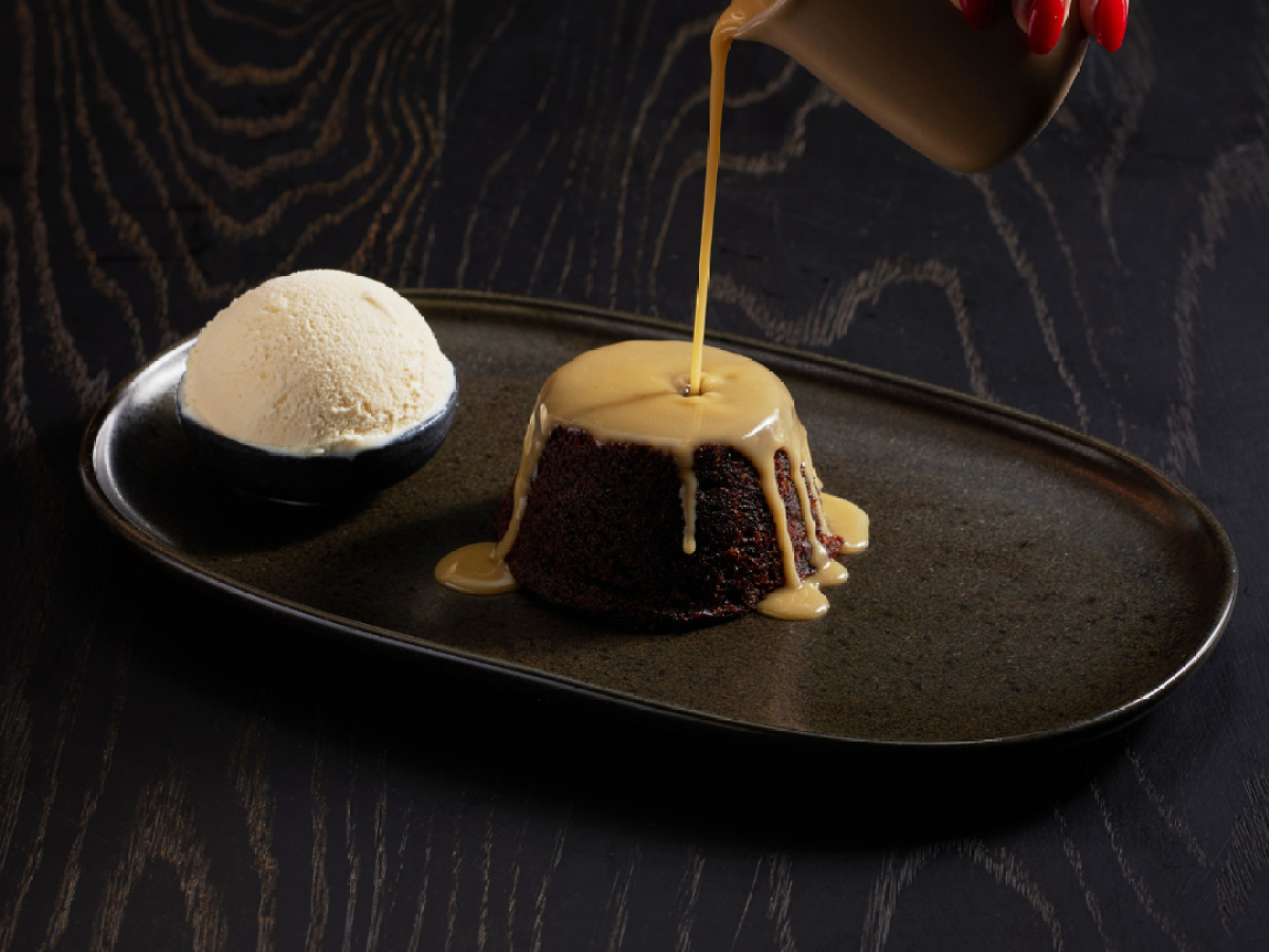 Hand pours sauce on cake with ice cream on dark plate.