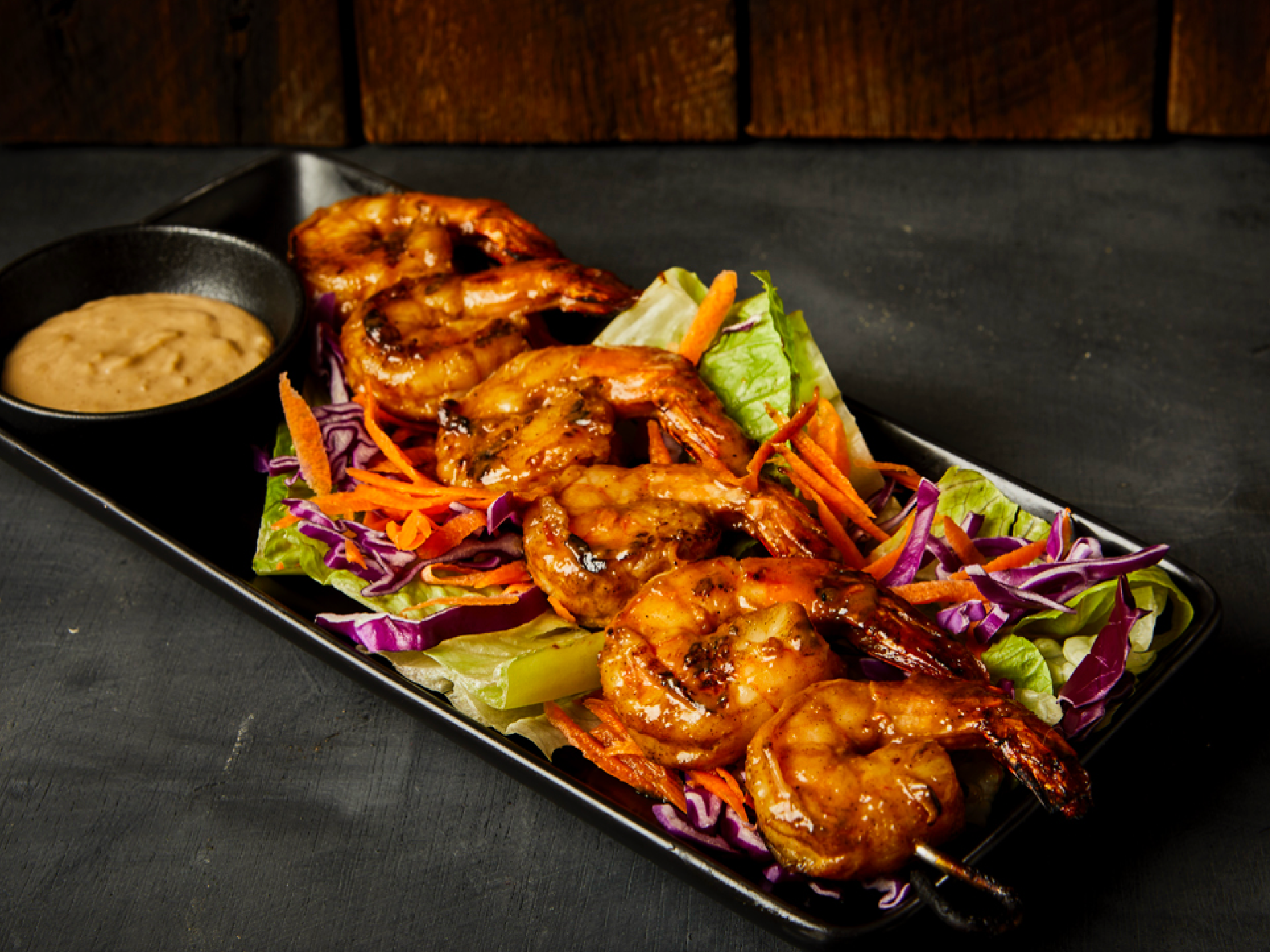 Grilled shrimp skewers on salad with dipping sauce on a black plate.