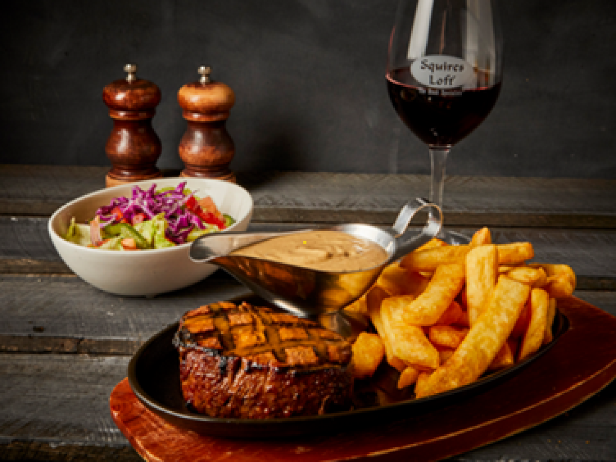 Steak with fries, salad, sauce, and a glass of red wine on a wooden table.