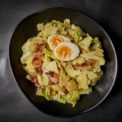 Caesar salad with boiled egg, bacon, and parmesan on a black plate.
