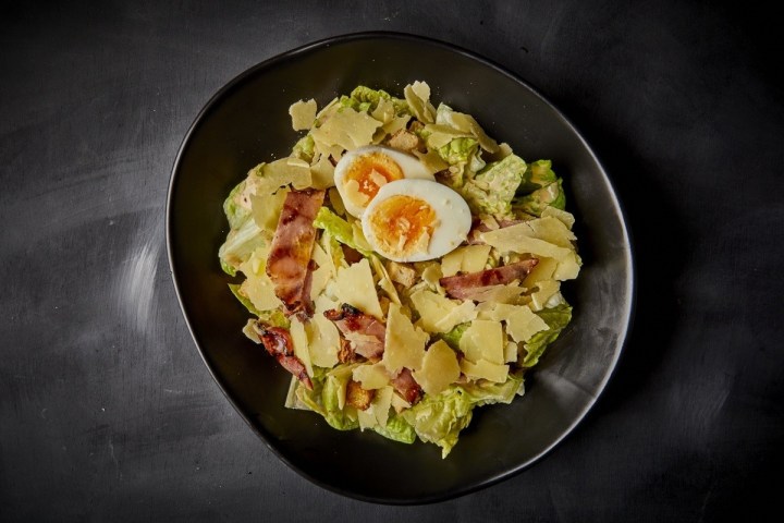 Caesar salad with boiled egg, bacon, and parmesan on a black plate.